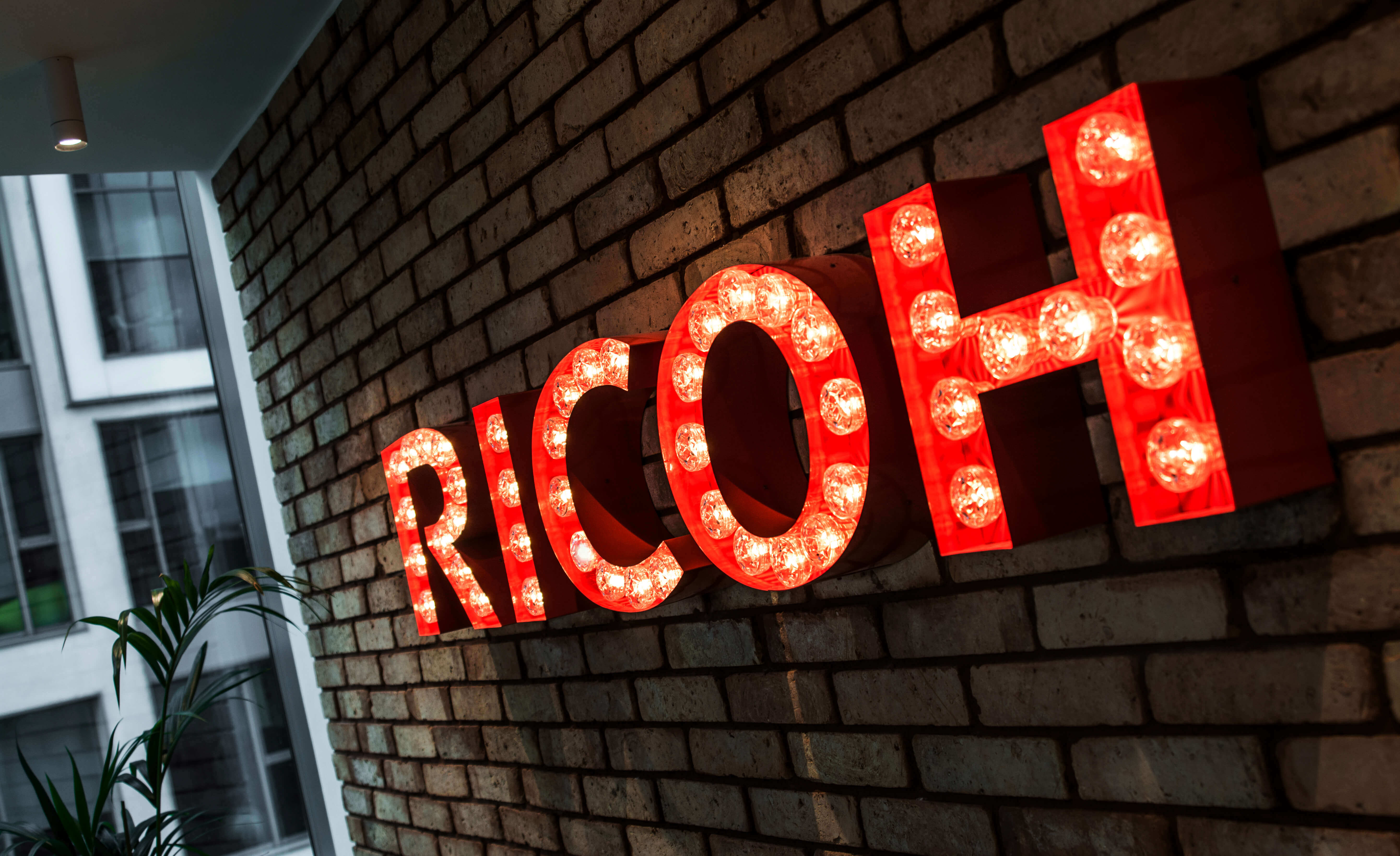 RICOH - Buchstaben mit Glühbirnen auf einer Backsteinmauer